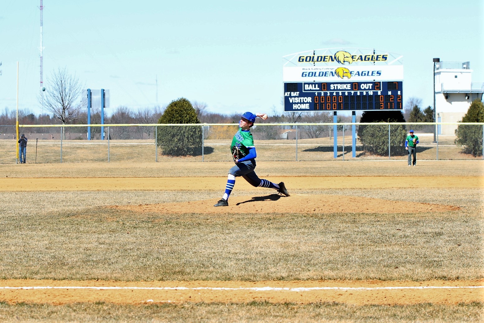 Connor Olson about to release a pitch, Rock Valley scoreboard in the background