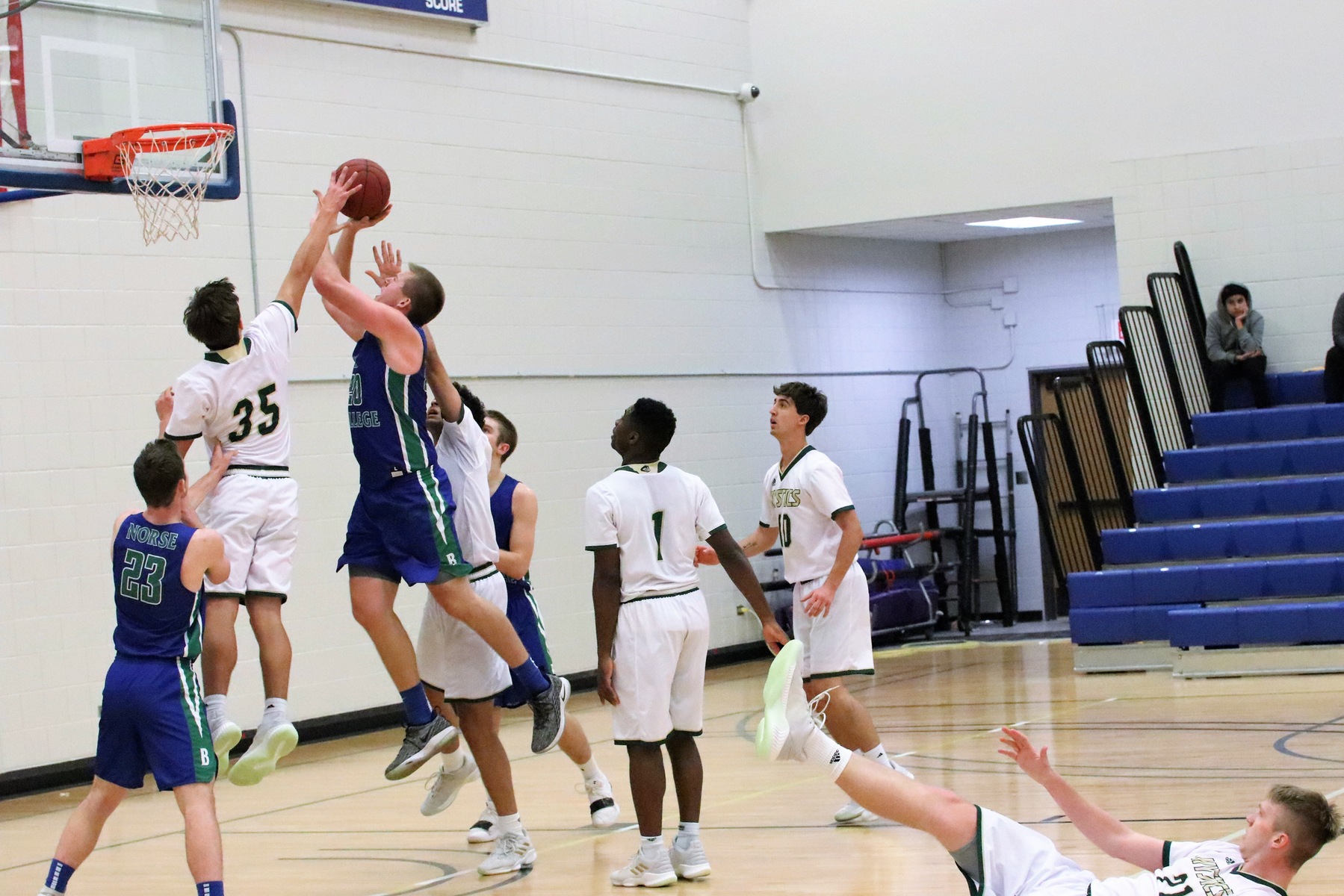 Logan Hardwick in mid air about to shoot near the basket.  Two defenders are closely defending him, one with an arm extended toward the ball.  