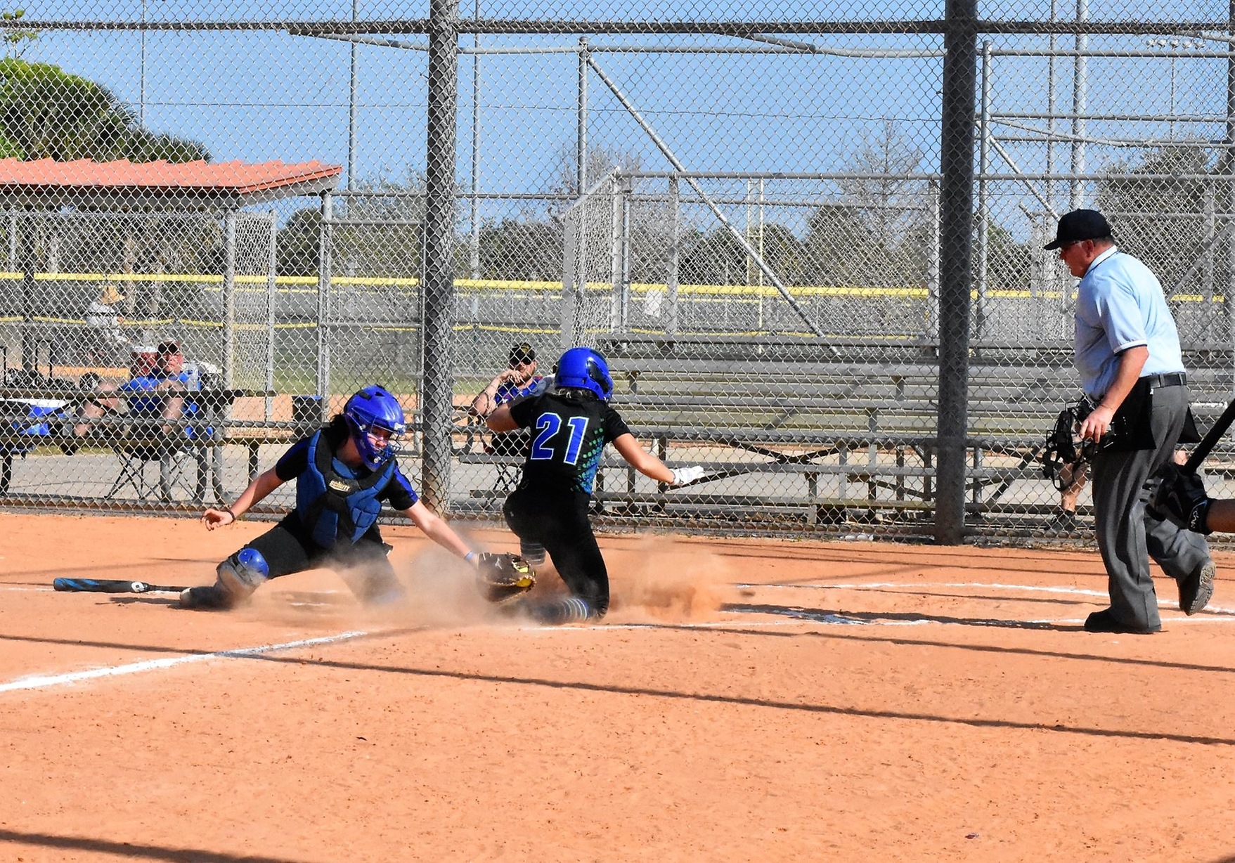 Emily Bruntjens after a slide into home plate. The catcher is reaching behind Emily with the ball in her glove.  The umpire looks on from foul territory